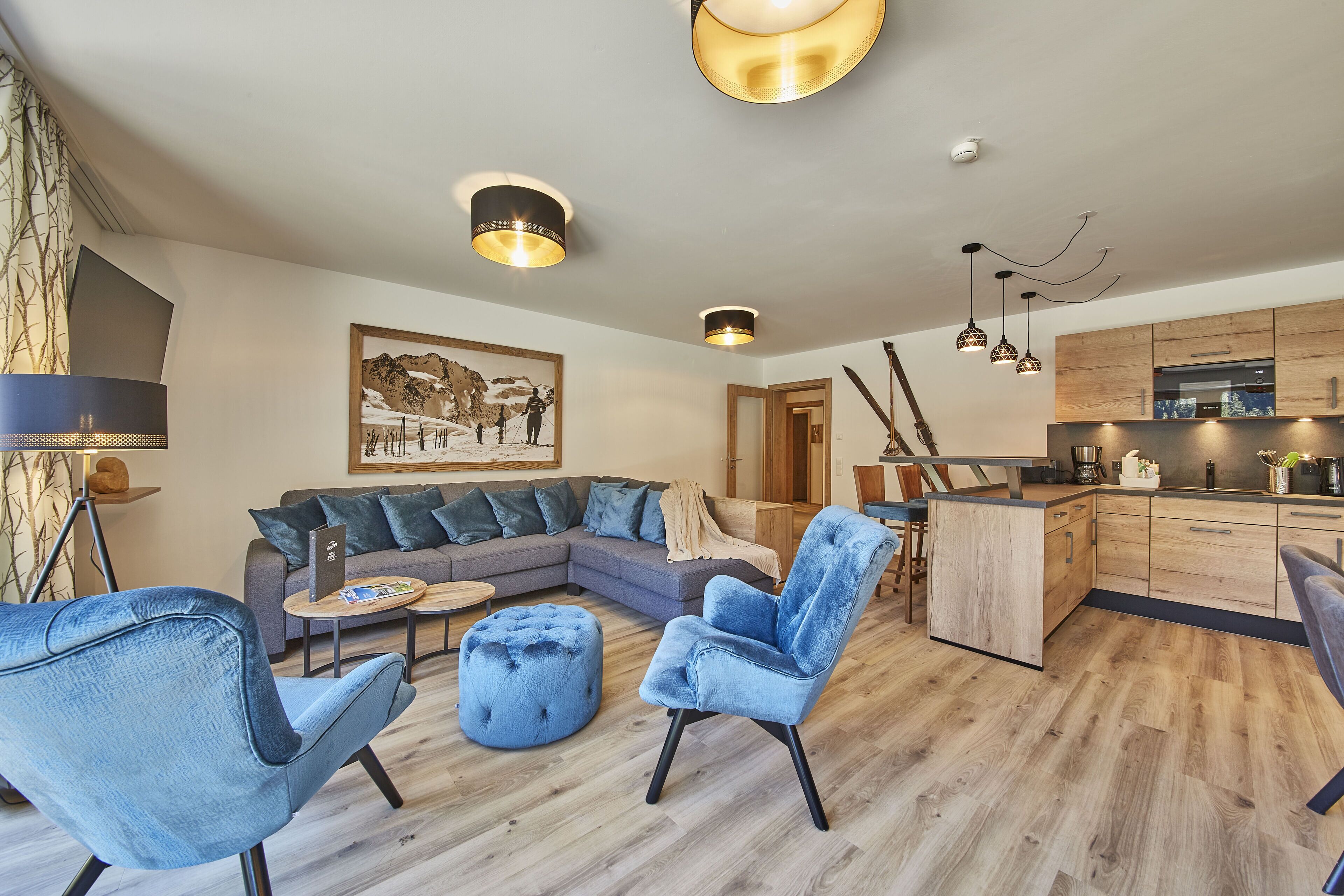 A modern living room with a sectional sofa, blue armchairs, and a round ottoman. There is a kitchen area with wooden cabinets and a dining table.