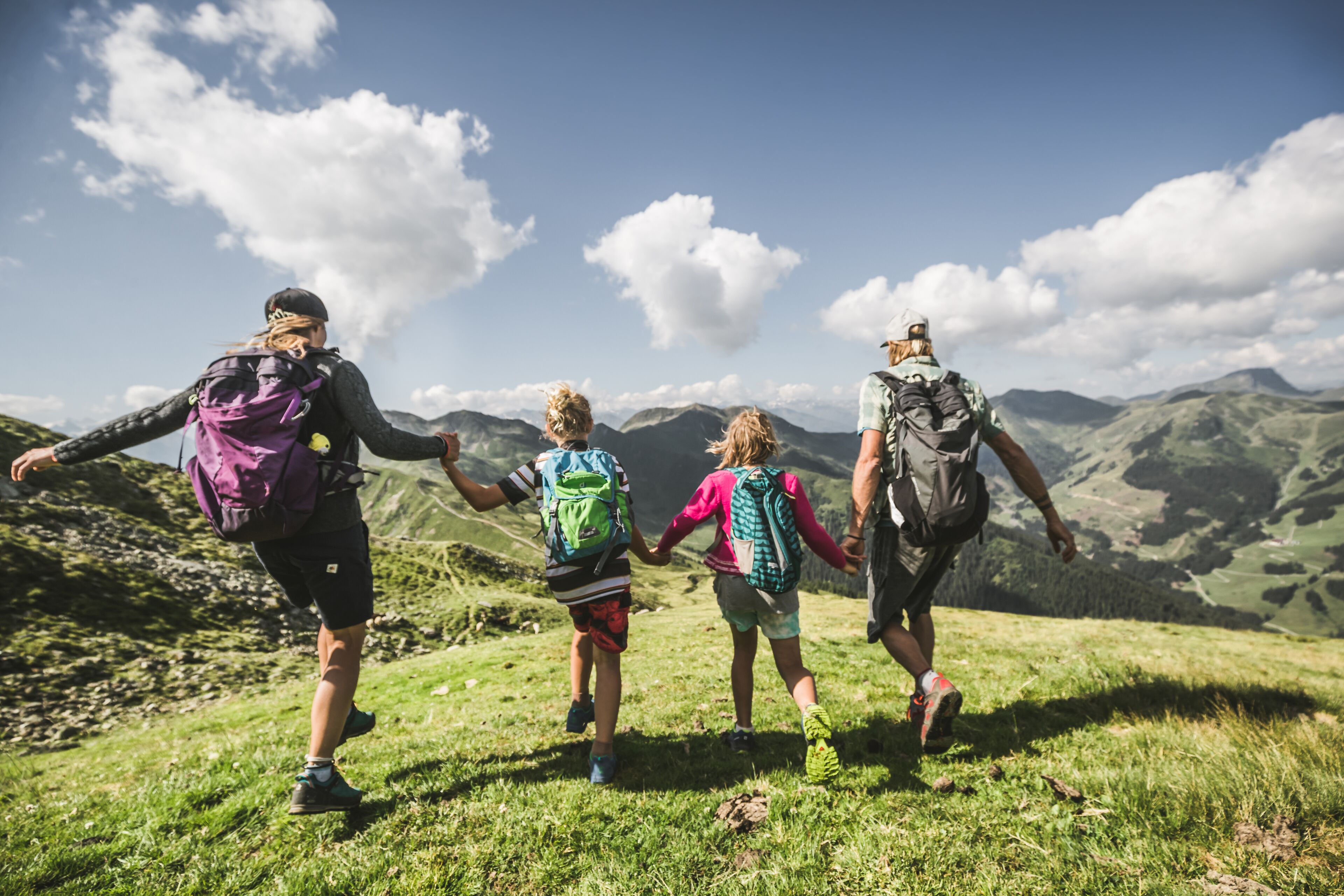Vier mensen wandelen op een bebost bergpad, met een heldere lucht en bergen in de verte.