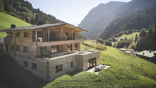 Uma casa moderna de madeira com dois andares e varanda, situada num vale montanhoso com vegetação exuberante e colinas ao longe.