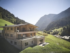 A modern two-story wooden house with a balcony, set in a mountainous valley with lush greenery and distant hills.