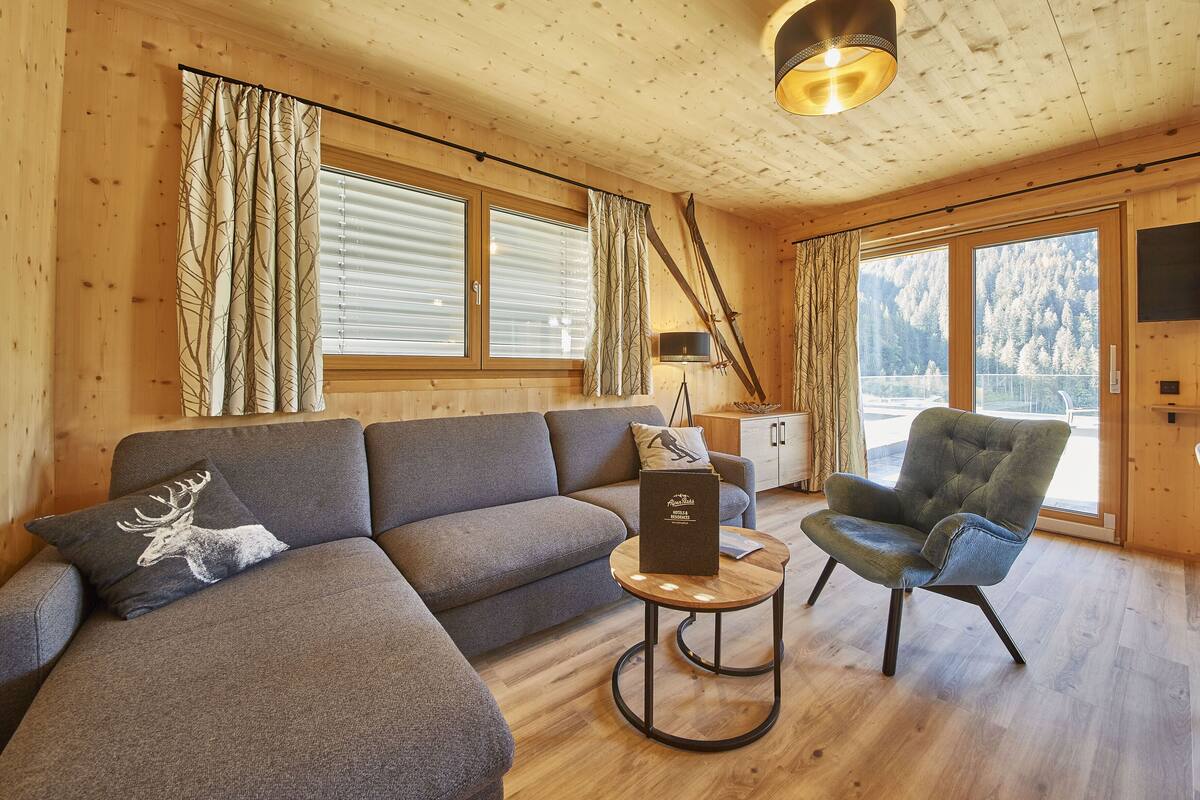 A cozy room with a sofa, armchair, and a small table. There are large windows with curtains, a lamp, and a view of trees outside.