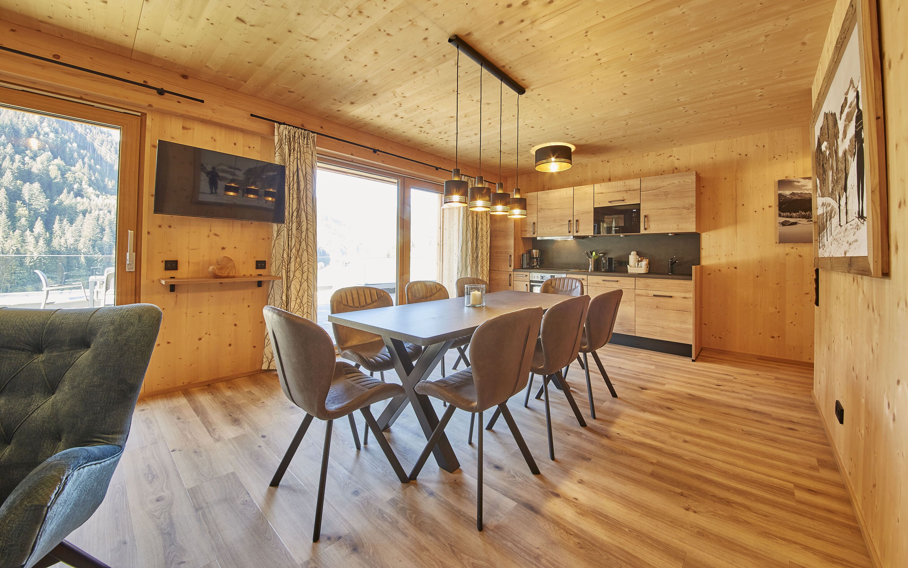 Un interior de madera con una mesa de comedor, sillas, una zona de cocina y un amplio ventanal con vistas a un área boscosa.