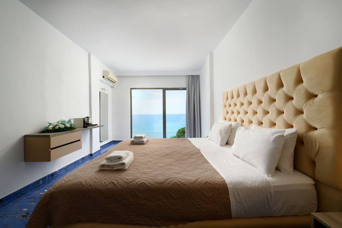Een hotelkamer met een groot bed, uitzicht op zee, een klein bureau met een plant en een raam met gordijnen.