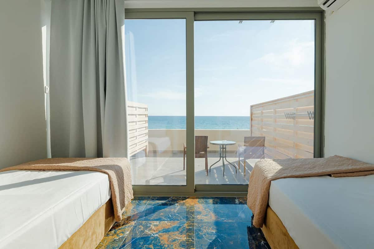 Een hotelkamer met twee bedden, uitzicht op zee en een balkon met een tafel en stoelen.