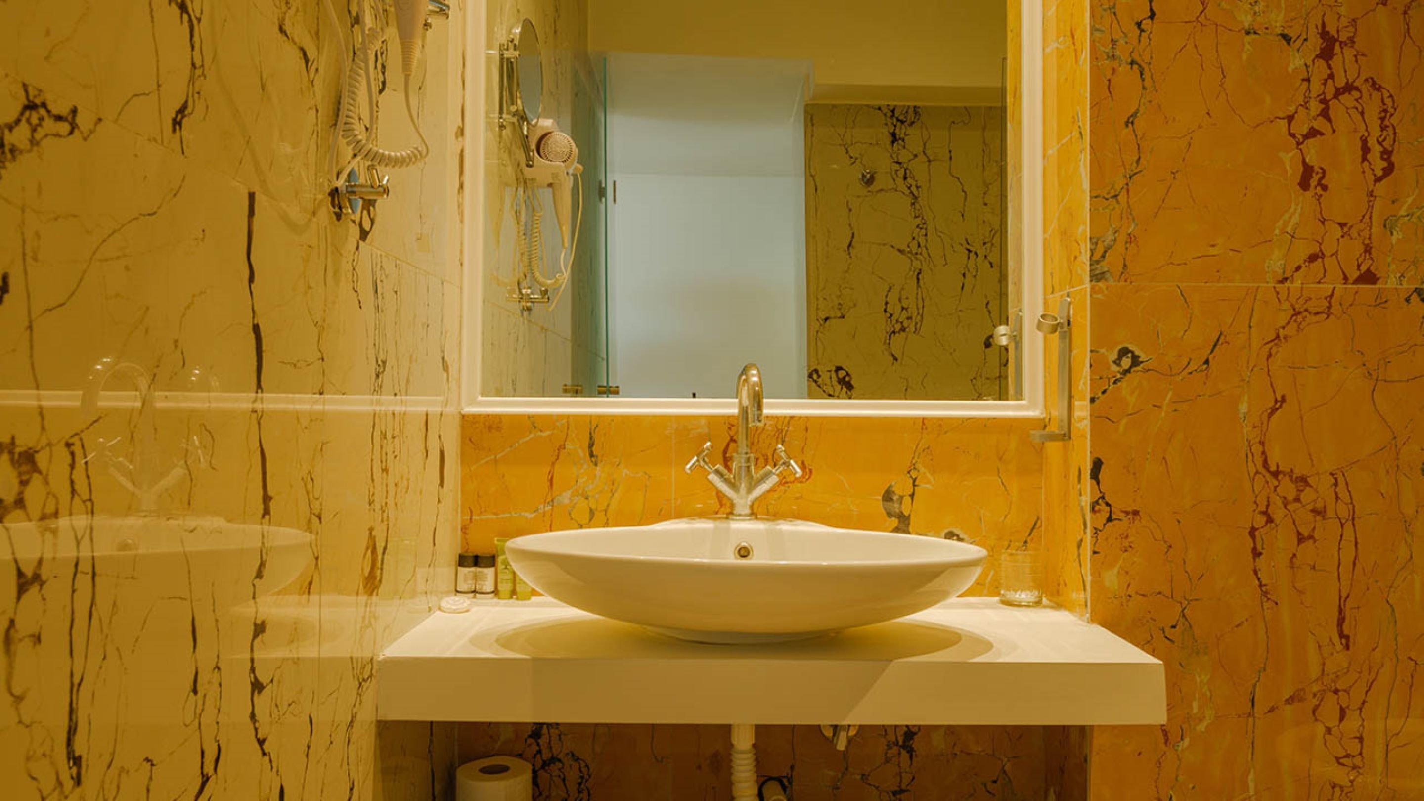Un baño con un lavabo blanco, un espejo y paredes de mármol amarillo.