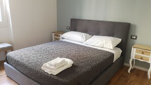 Ein ordentlich bezogenes Bett mit einem gemusterten Überwurf und weißen Kissen, ein Nachttisch mit Lampe sowie eine kleine Bank.