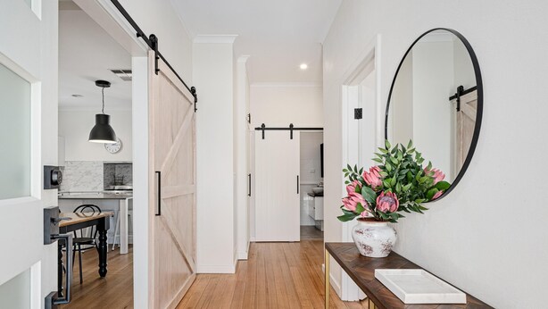 Un couloir avec un plancher en bois, une porte blanche, un miroir et un vase rempli de fleurs.