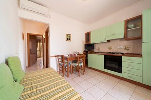 A compact kitchen with green cabinets, a dining table with chairs, and a sofa in the living area. - Villetta Grazia - 1 (Capoliveri)