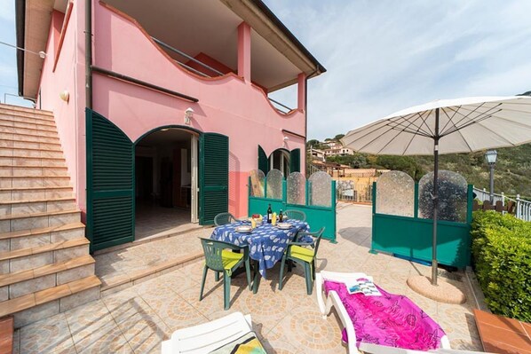 A pink building with a covered outdoor area, a table with chairs, and a large umbrella. - Villetta Grazia 3 - Capoliveri (Capoliveri)
