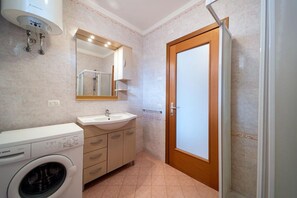 A compact bathroom with a washing machine, sink, mirror, and a door.