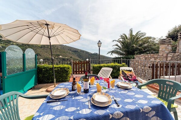 An outdoor dining area with a table set for two, a large umbrella, and a scenic view of hills.