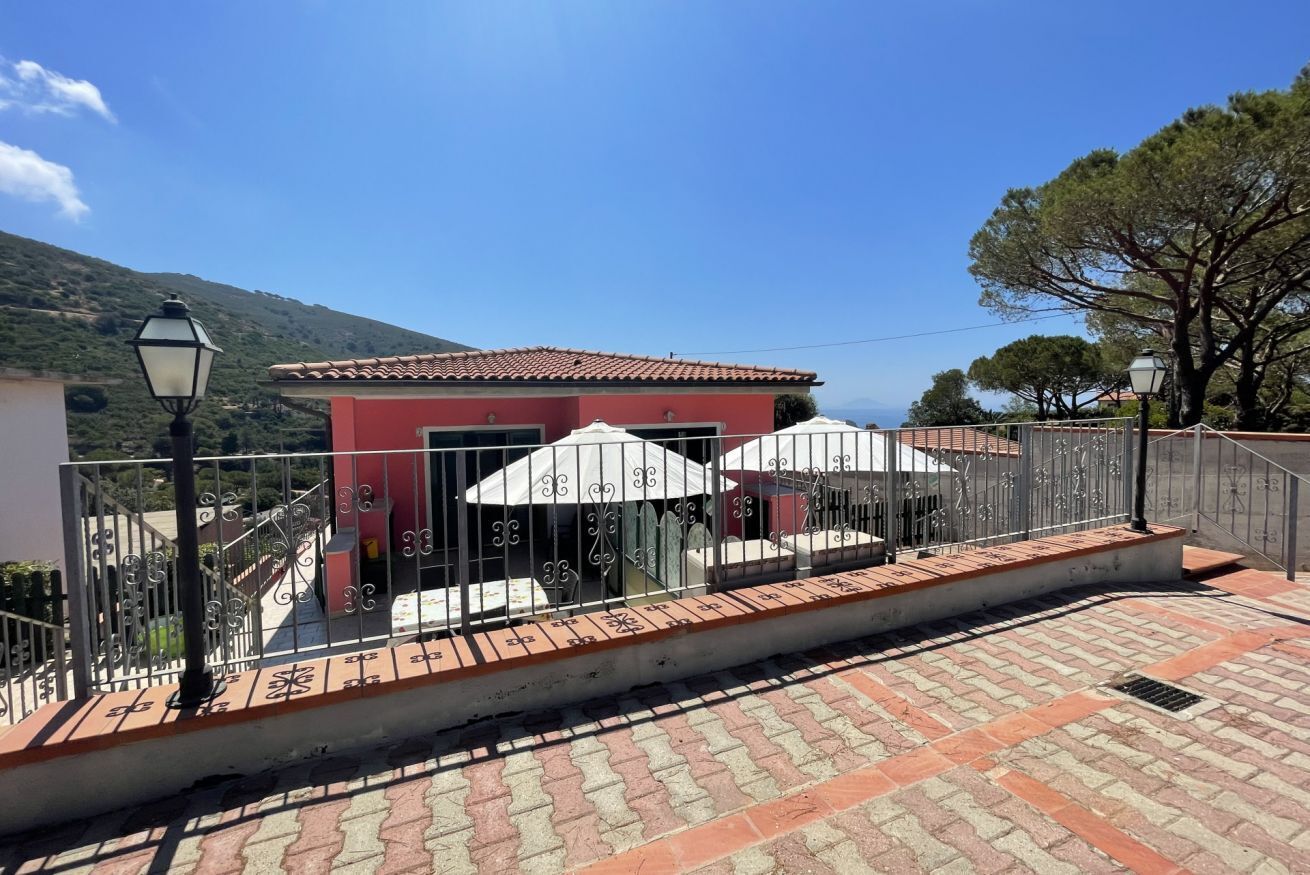 A red building with a tiled roof, a paved terrace with a metal railing, and a view of the sea.