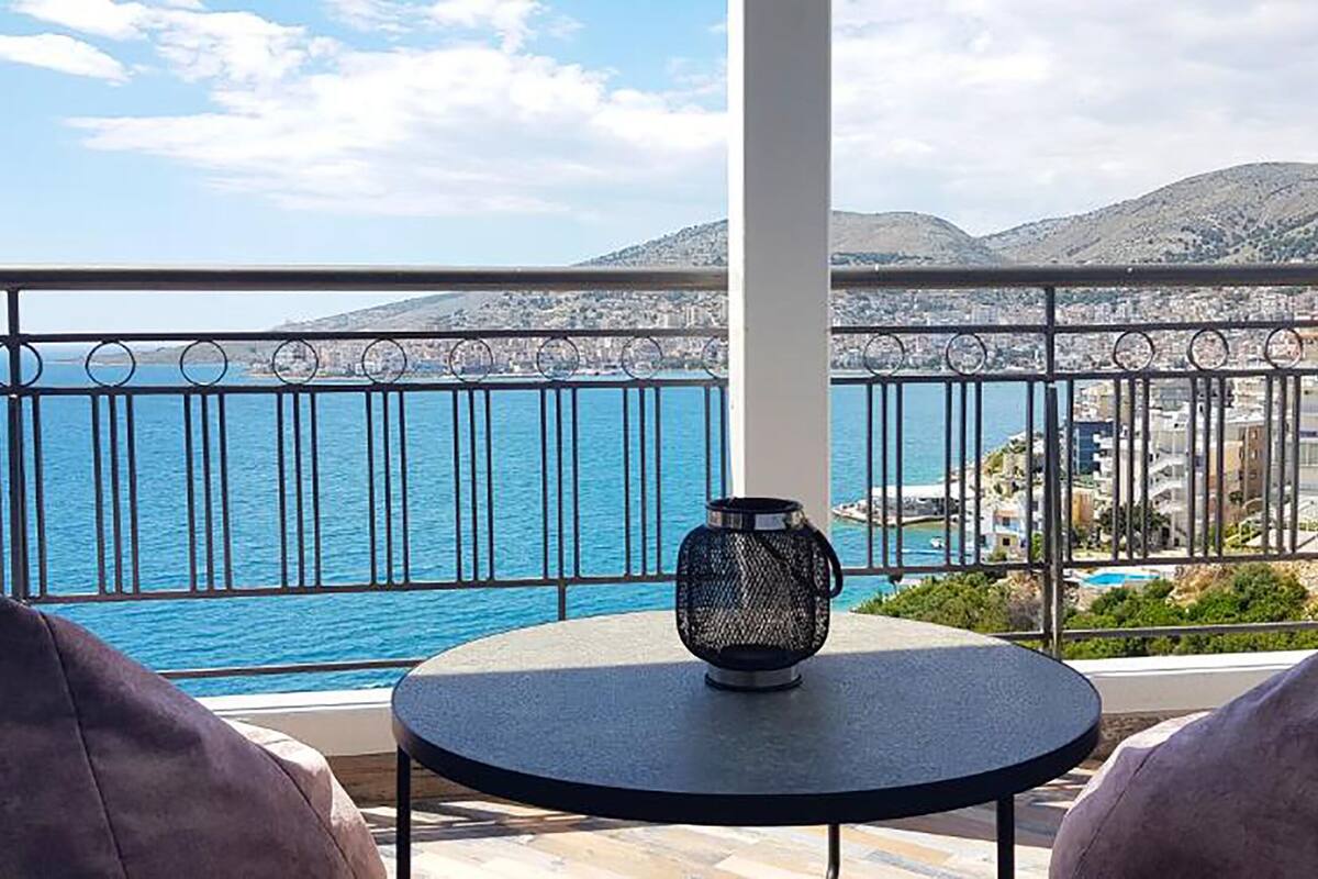 Un balcó amb una taula rodona i un vas, amb vista a una ciutat costanera i una visió clara de la mar i les muntanyes llunyanes.