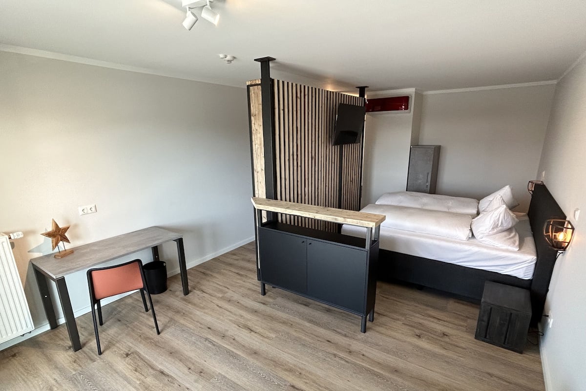 A modern hotel room with a wooden desk, a bed with white linens, a black headboard, and a small nightstand with a lamp.