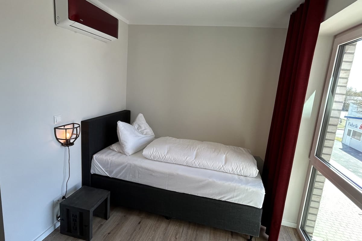 A single bed with white bedding in a room with a window, a wall-mounted light, and a small table.