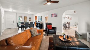 A spacious living area with a leather sofa, a black armchair, and a dining table with chairs. A ceiling fan is visible, and the room has a modern, clean design with white walls and a tiled floor. - This spacious home has all you need with 2 primary suites, 2 guest bedrooms, and 3 bathrooms. There is no shortage of living space with a living room, 2 dining rooms, open floor plan w/fully-stocked kitchen, and onsite laundry. (El Paso)