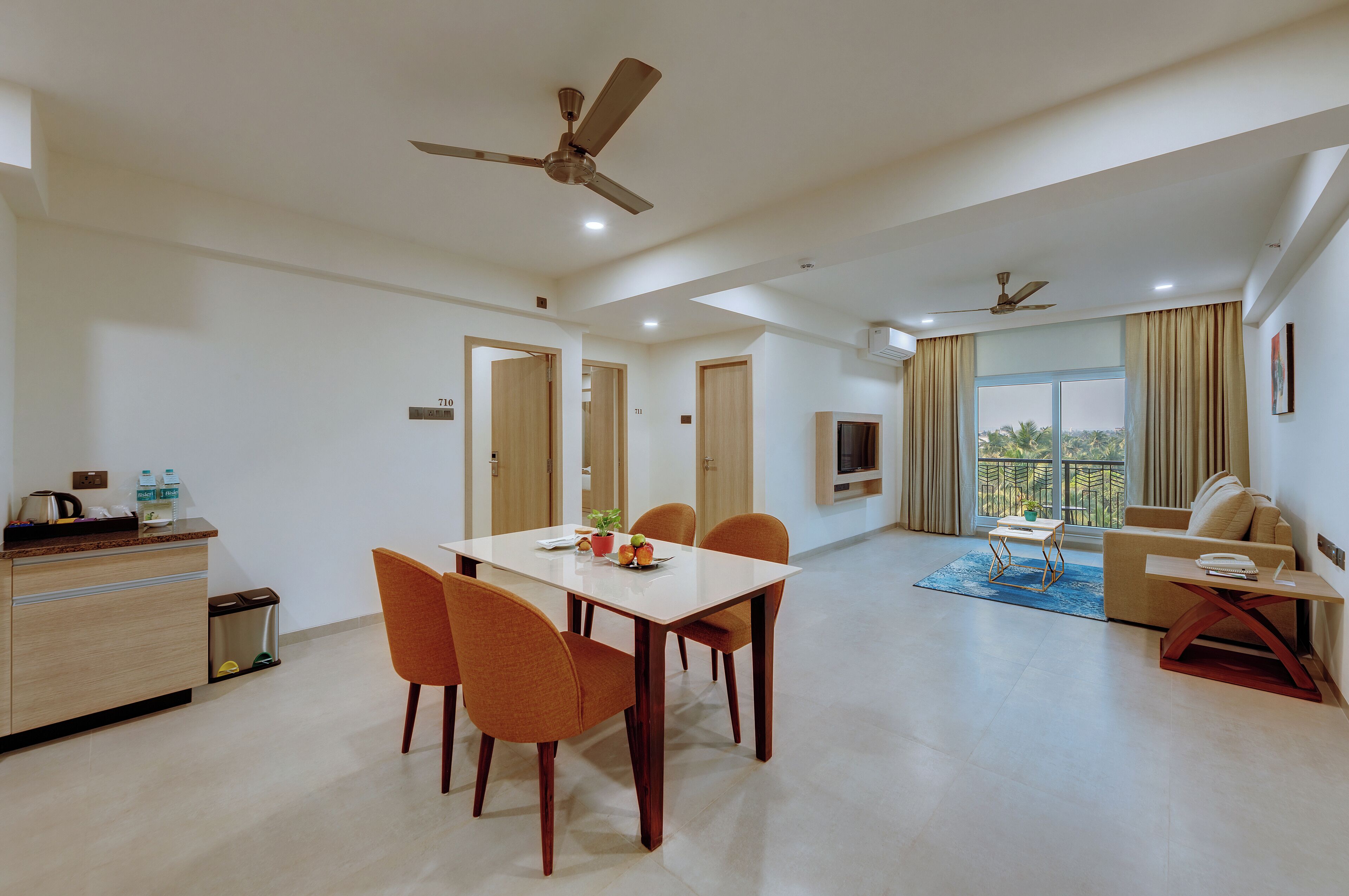 A modern living room with a dining area, a sofa, and a TV. There are ceiling fans, a window with curtains, and a balcony view.