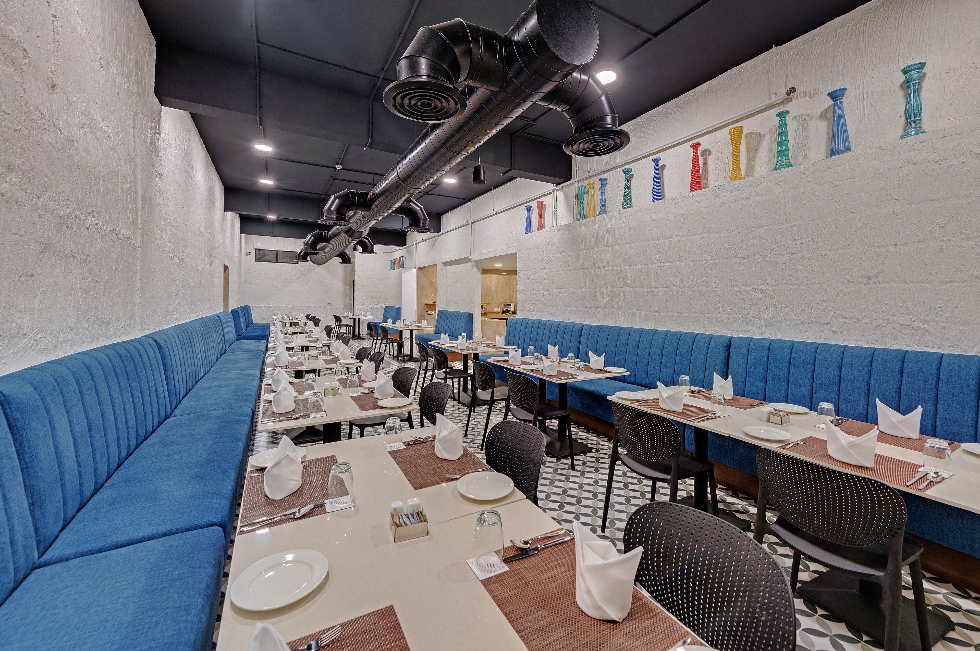 A restaurant with blue booths, white walls, and black ceiling ducts. Tables are set with white tablecloths and silverware.