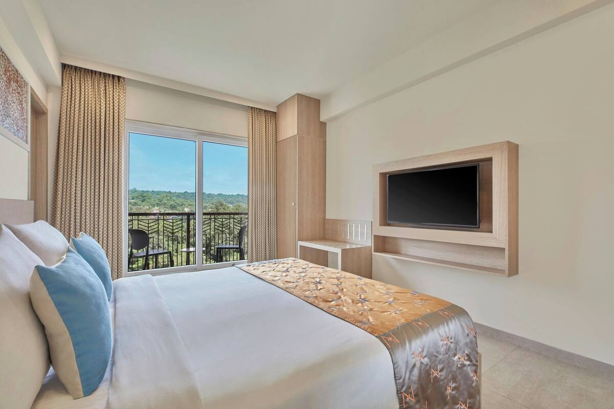 A hotel room with a large bed, a TV mounted on the wall, and a balcony with a view of greenery.