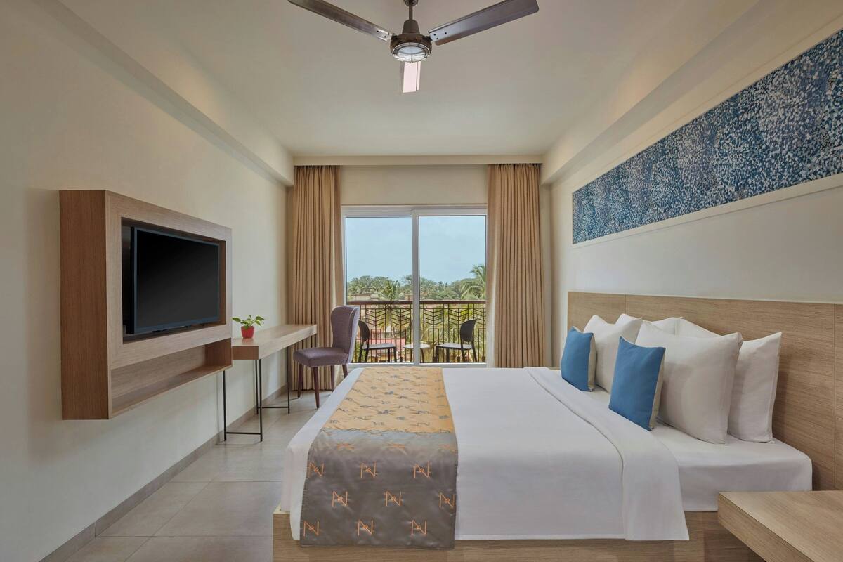 A hotel room with a bed, a TV mounted on the wall, a desk with a chair, a balcony with a table and chairs, and a ceiling fan.