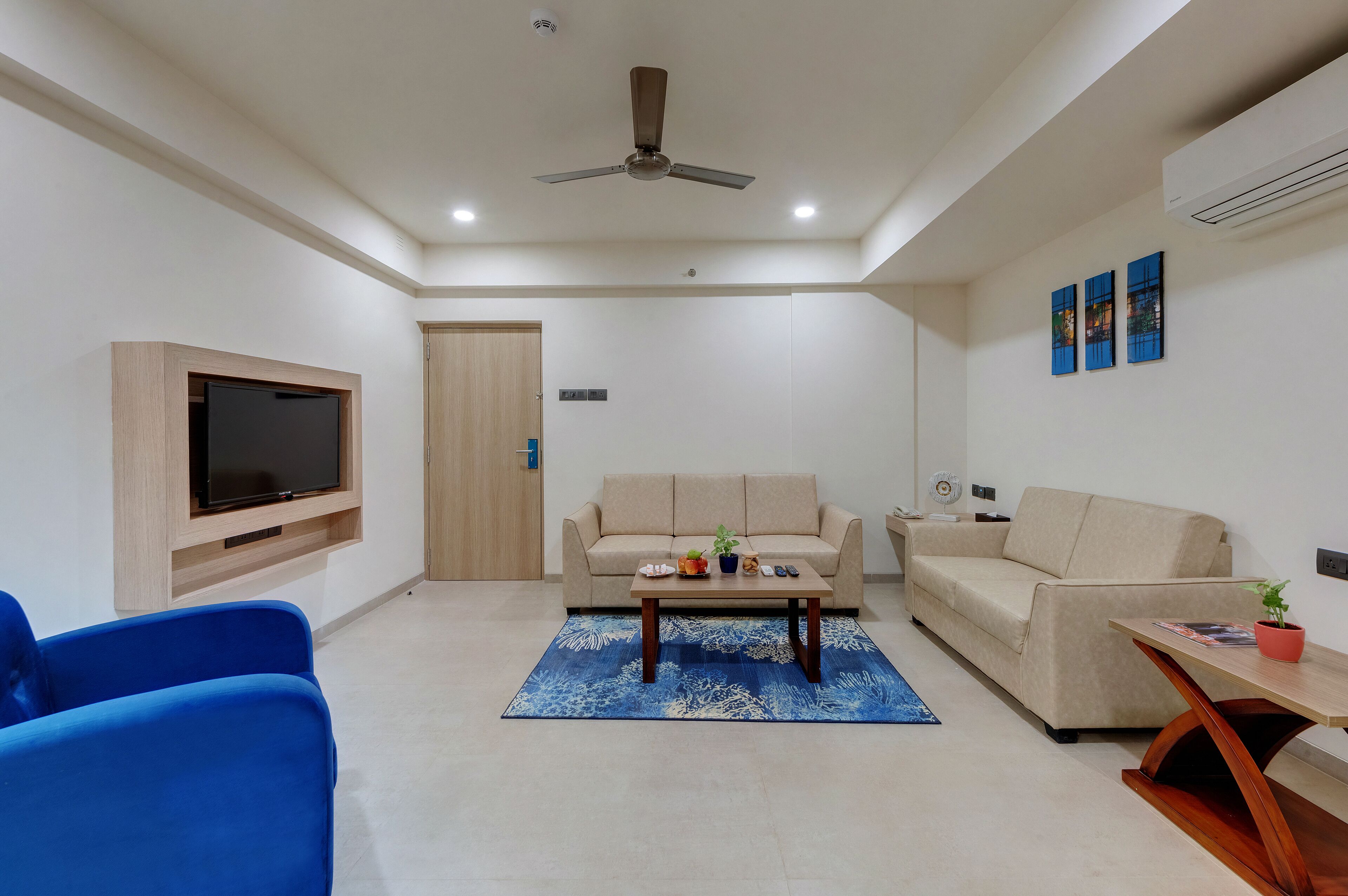 Ein modernes Wohnzimmer mit Flachbildfernseher an der Wand, einem beigen Sofa, einem blauen Sessel, einem Couchtisch und einem Deckenventilator.