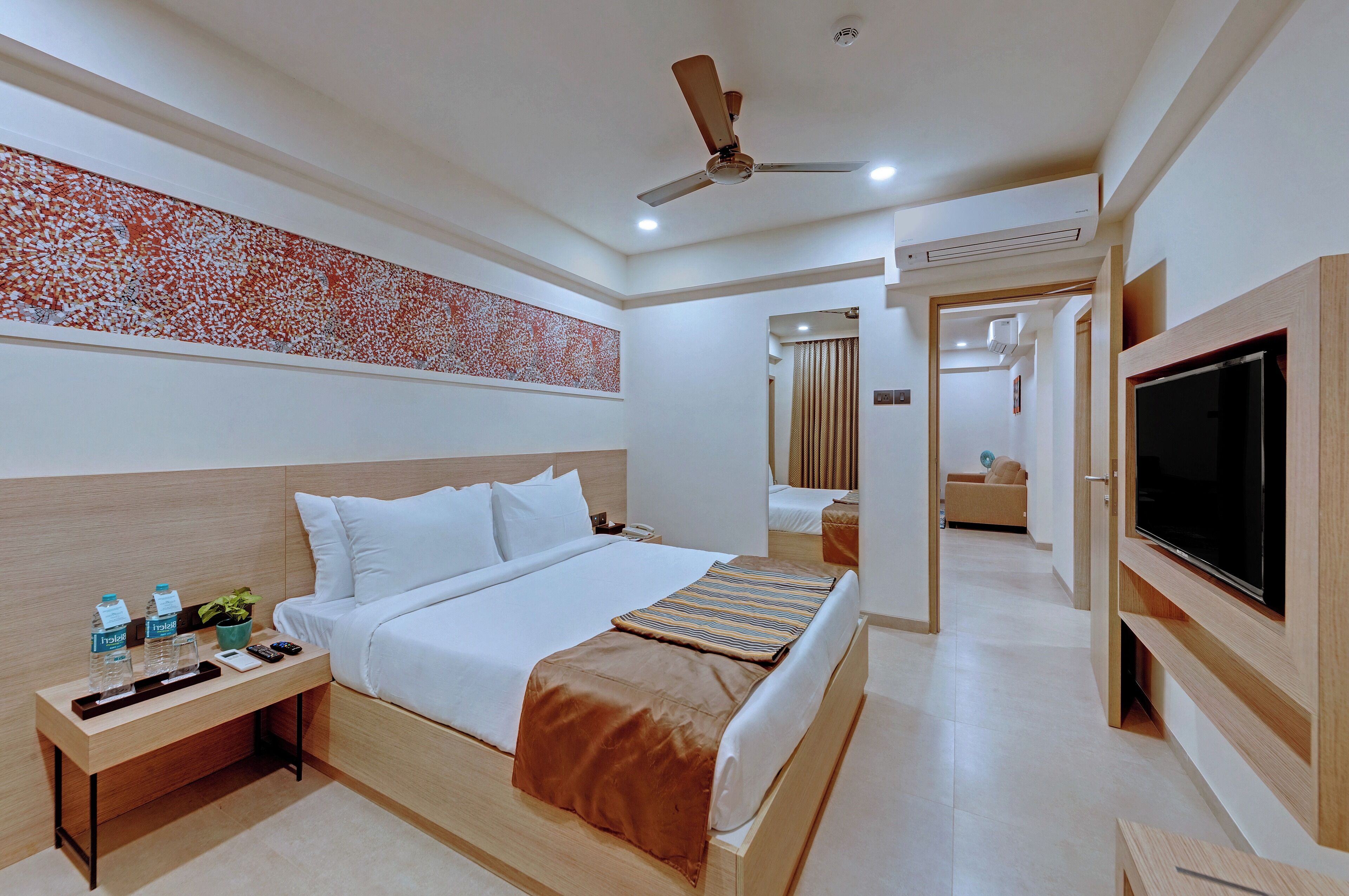 A hotel room with a bed, bedside table, TV, ceiling fan, and a decorative wall piece.