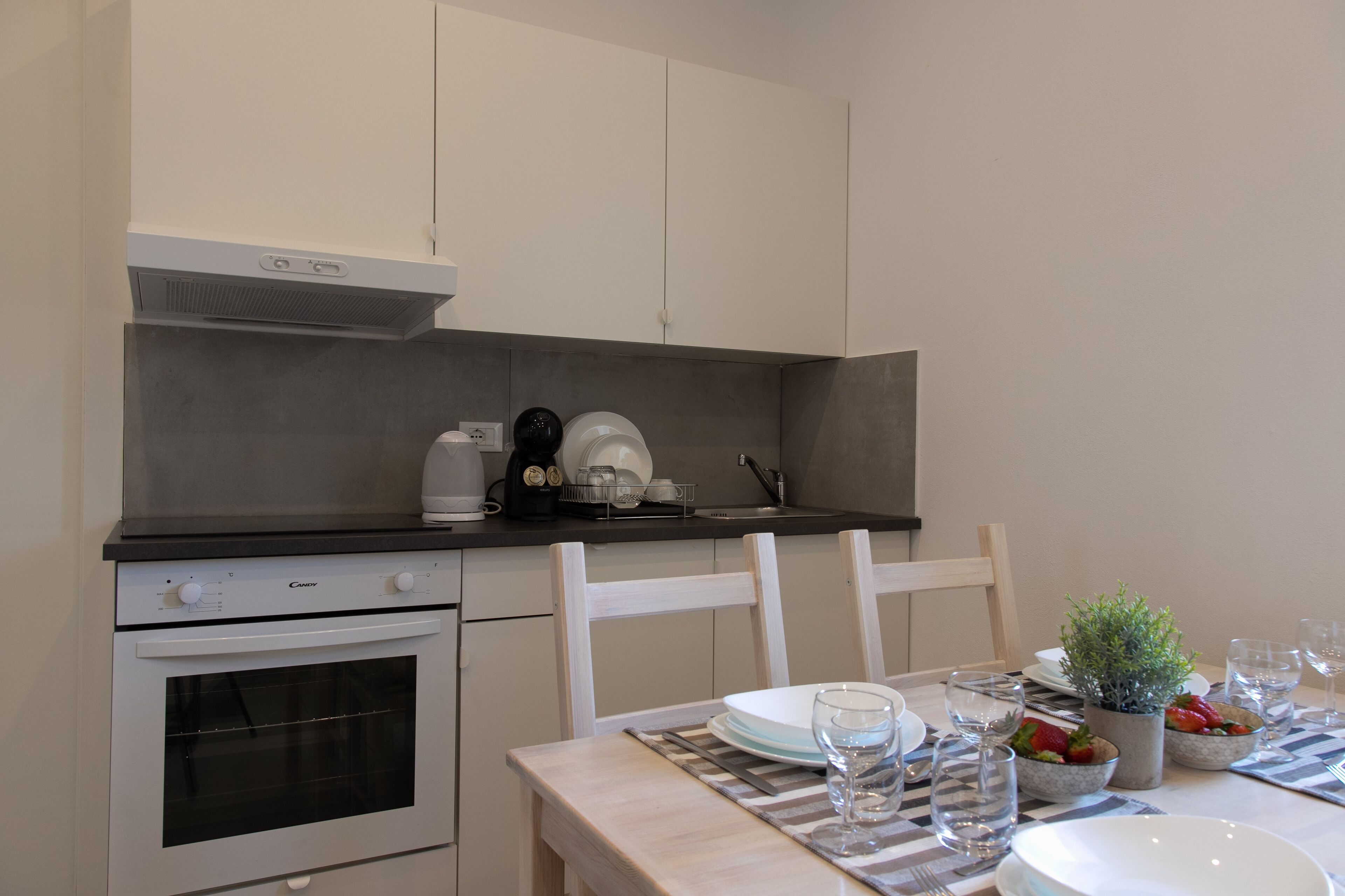 Moderni keittiö, jossa on valkoiset kaapit, sisäänrakennettu uuni ja musta työtaso. Ruokapöytä on katettu lautasilla, laseilla ja siinä on pieni ruukkukasvi.
