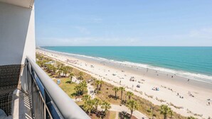 Un balcon donnant sur une plage, avec une vue dégagée sur l’océan et les personnes se promenant sur la plage.