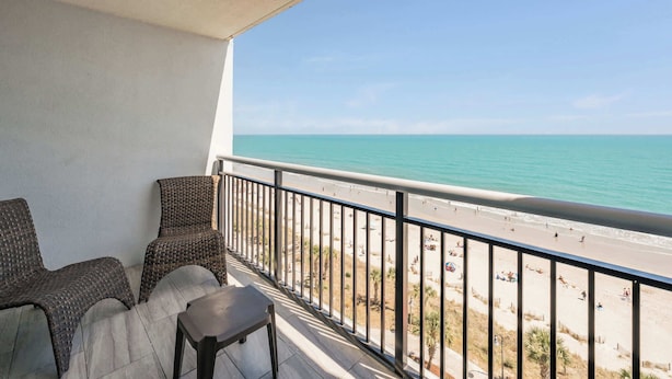 Un balcon avec des fauteuils en osier et une table, donnant sur une plage et offrant une vue dégagée sur l’océan.