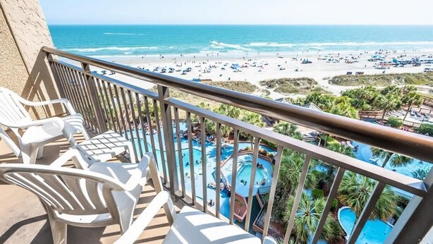 Ein Balkon mit weißen Plastikstühlen mit Blick auf einen Strand und einen Poolbereich mit mehreren Schwimmbecken.
