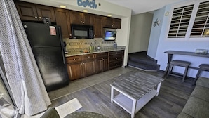 Eine kompakte Küche mit dunklen Holzschränken, einem schwarzen Kühlschrank und einer Mikrowelle. Es gibt eine kleine Kücheninsel mit einem Spülbecken und einem darüber montierten Fernseher.