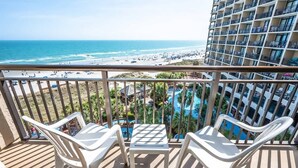 Ein Balkon mit weißen Plastikstühlen und einem kleinen Tisch, mit Blick auf einen Strand mit Pool und einem Resortgebäude.