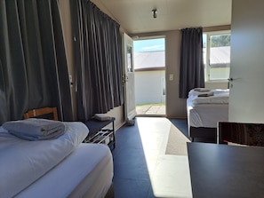 A hotel room with two beds, a small table, and a bench. There is a door leading to an outdoor area.