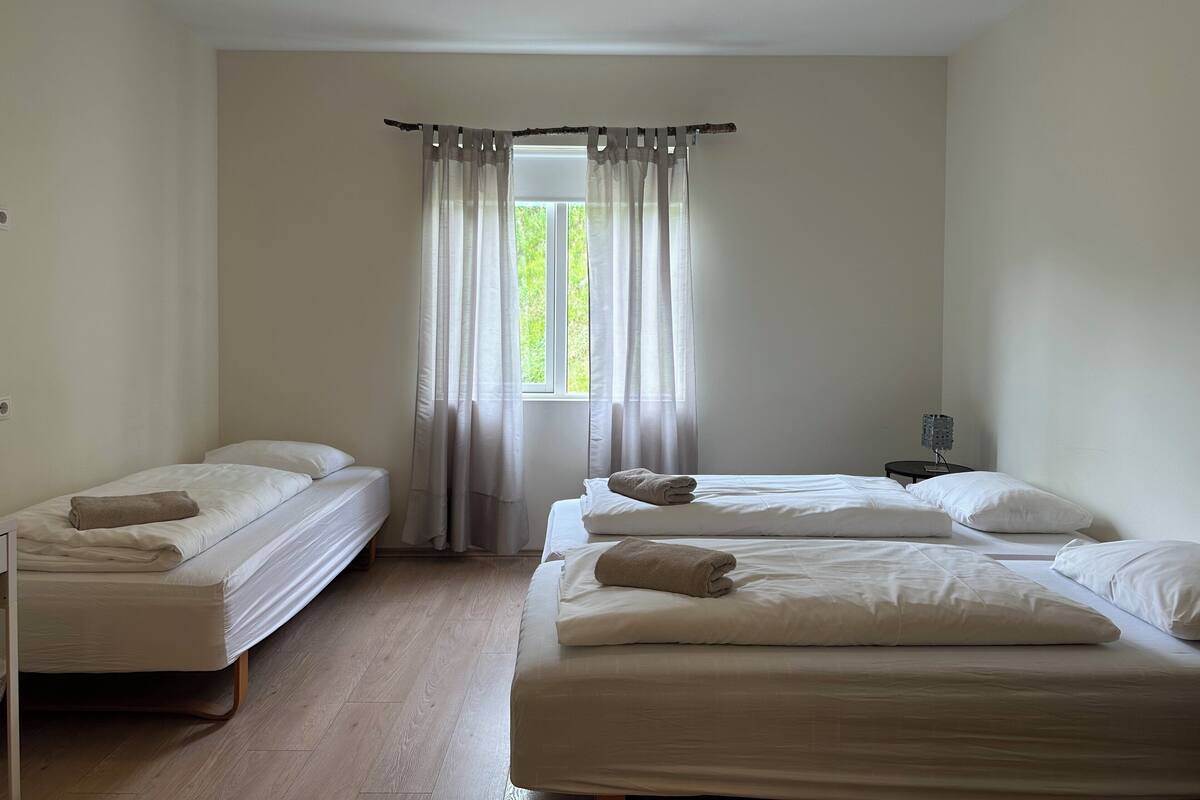 Habitación de hotel con dos camas individuales y una cama doble, una ventana con cortinas traslúcidas, una mesita de noche con lámpara y suelo de madera.