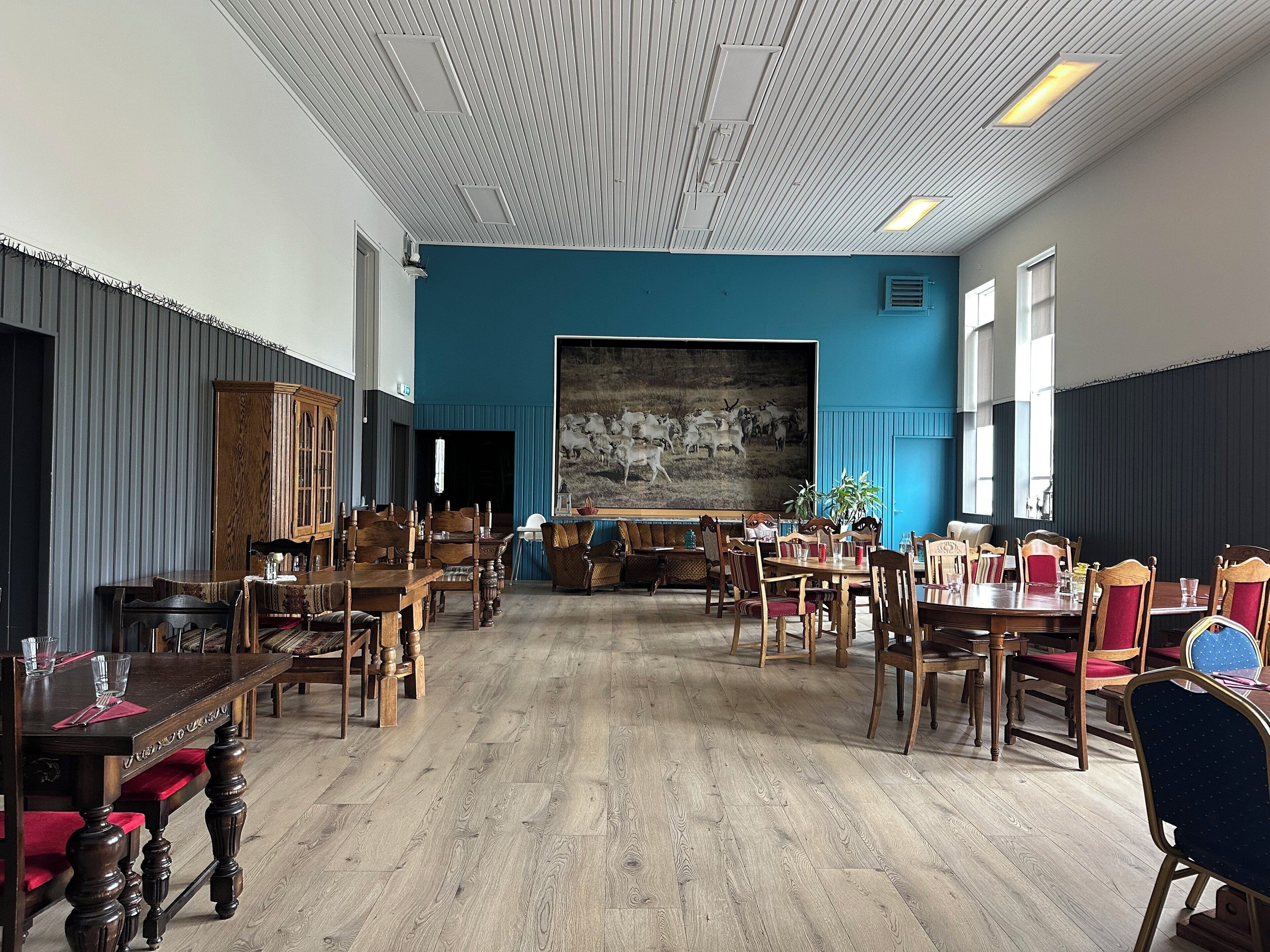 A spacious room with wooden tables and chairs, a large painting on the wall, and a blue accent wall.