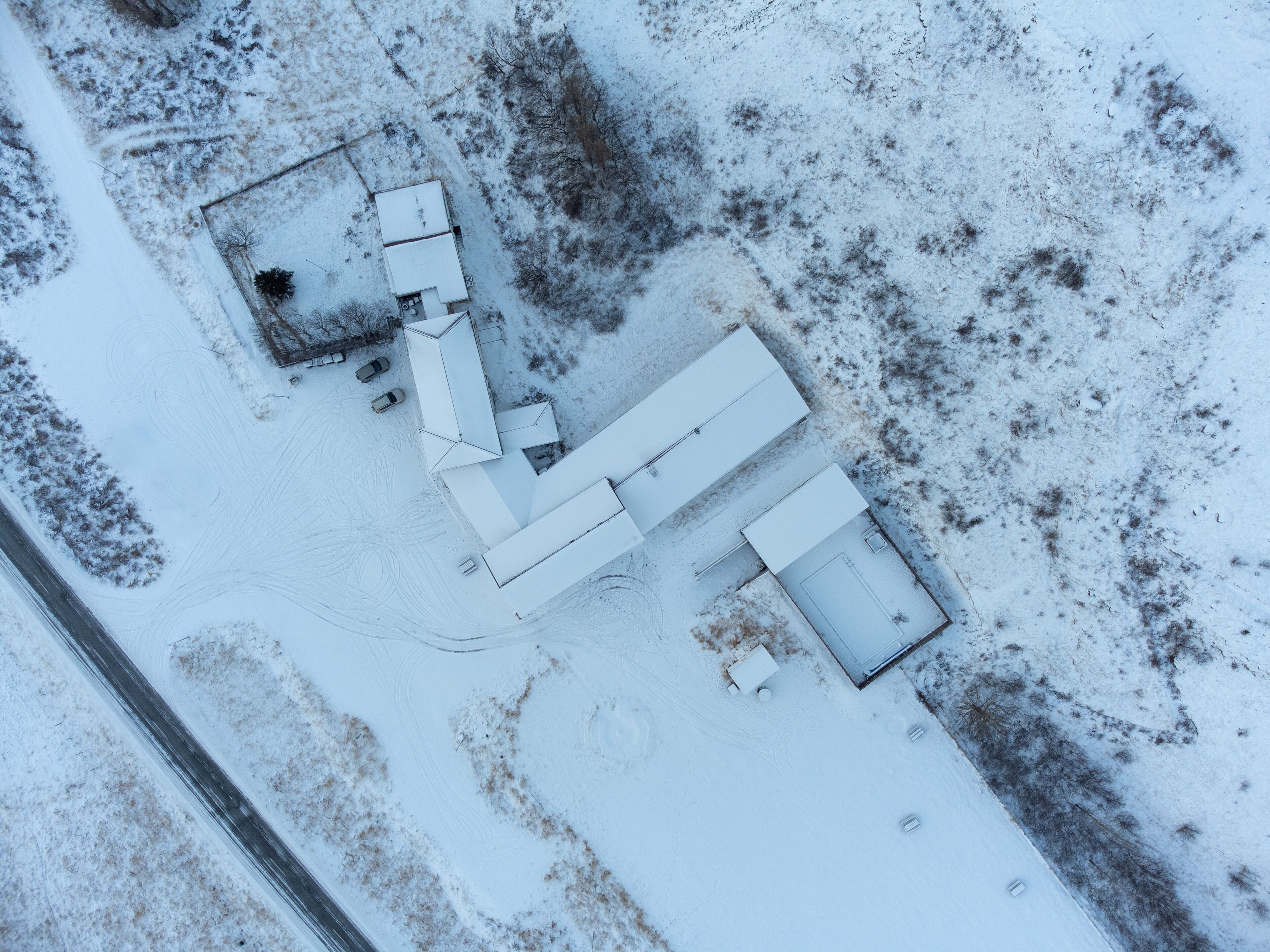 An aerial view of a snow-covered property with a large building, several smaller structures, and a paved driveway.