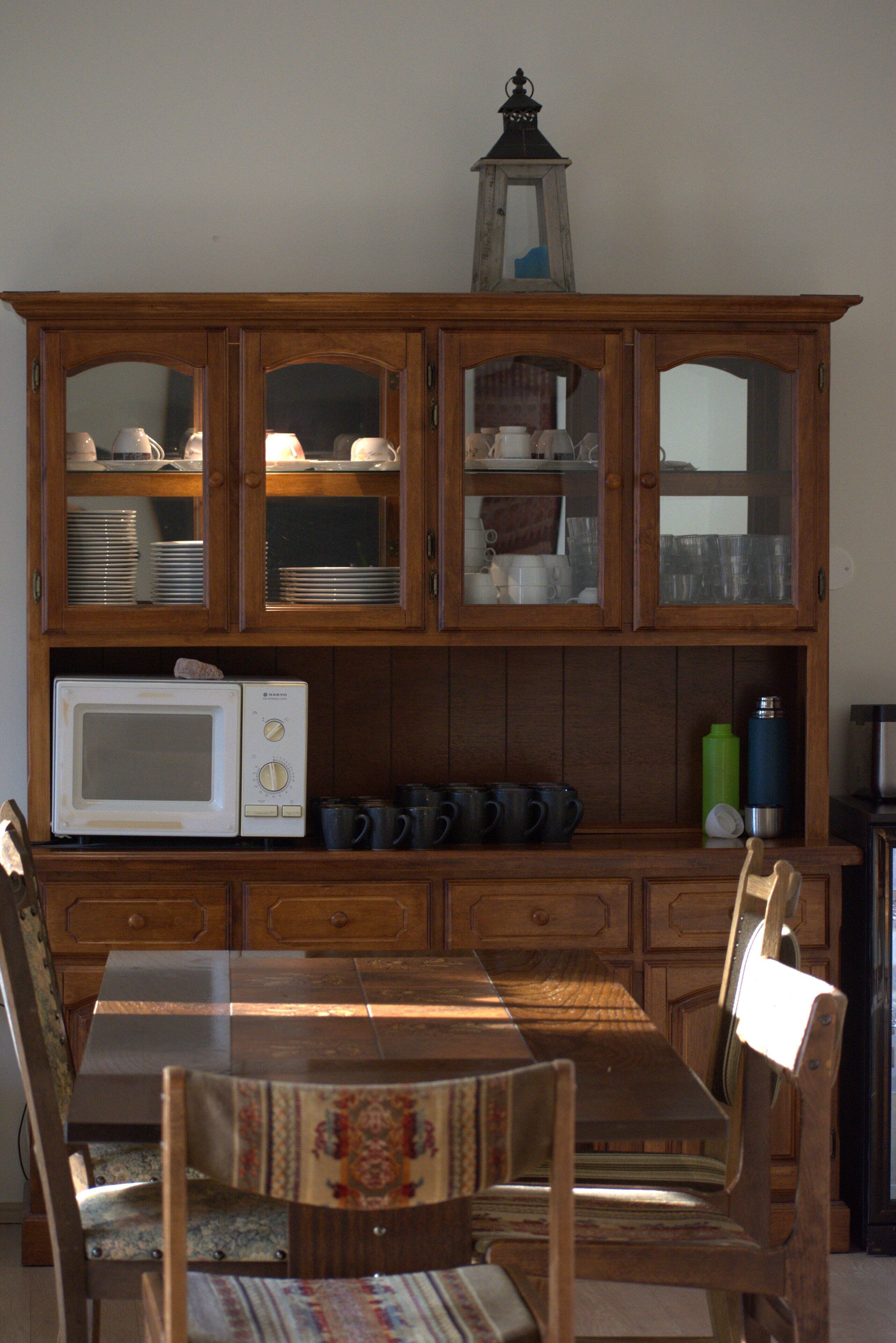 A dining area with a wooden table and chairs, a microwave, and a cabinet with glass doors containing dishes and cups.