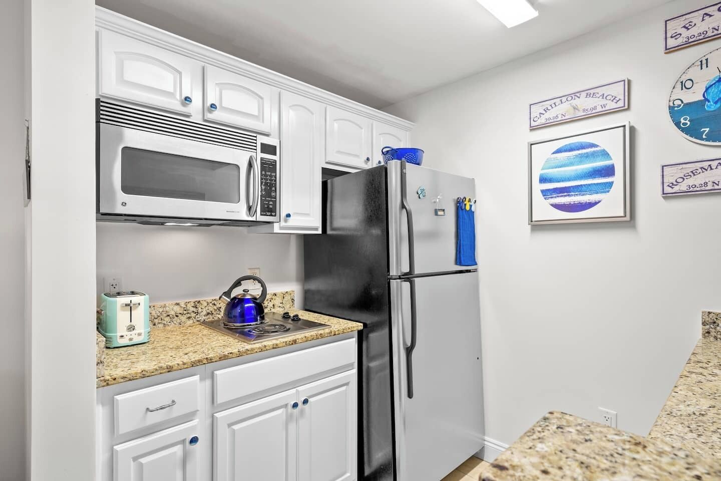 A kitchen with white cabinets, a stainless steel refrigerator, a microwave, and a blue kettle on the counter.