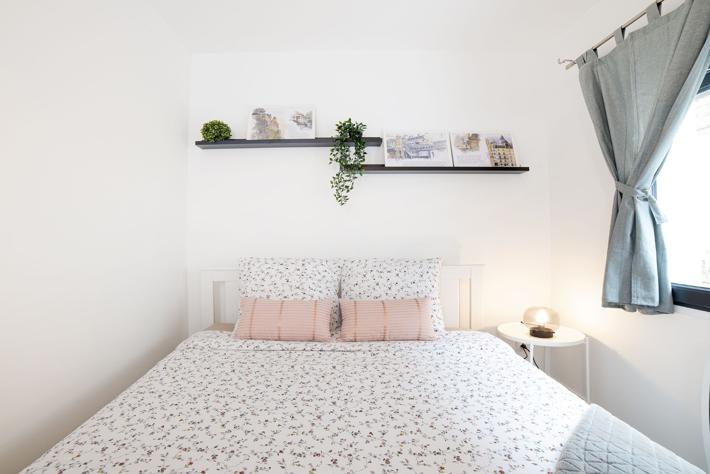 A neatly arranged bedroom with a bed, a bedside table with a lamp, a wall-mounted shelf with pictures and a plant, and a window with curtains.