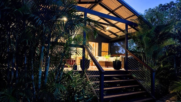 Una casa moderna con una scala in legno che conduce a un portico coperto, immersa in una fitta vegetazione tropicale.