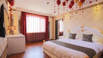 Ein Hotelzimmer mit einem großen Bett, einem Nachttisch, einer Lampe und Blick durch ein Fenster mit roten Vorhängen nach draußen.
