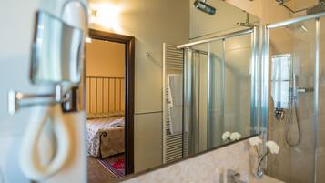 A modern bathroom with a glass shower enclosure, a large mirror, and a visible bed in the adjacent room.