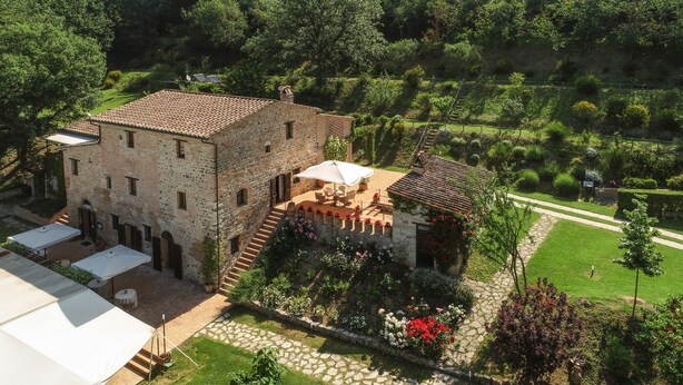 Un bâtiment en pierre avec un toit recouvert de tuiles, un escalier menant à un patio abrité et un jardin agrémenté de plantes et de fleurs en pot.