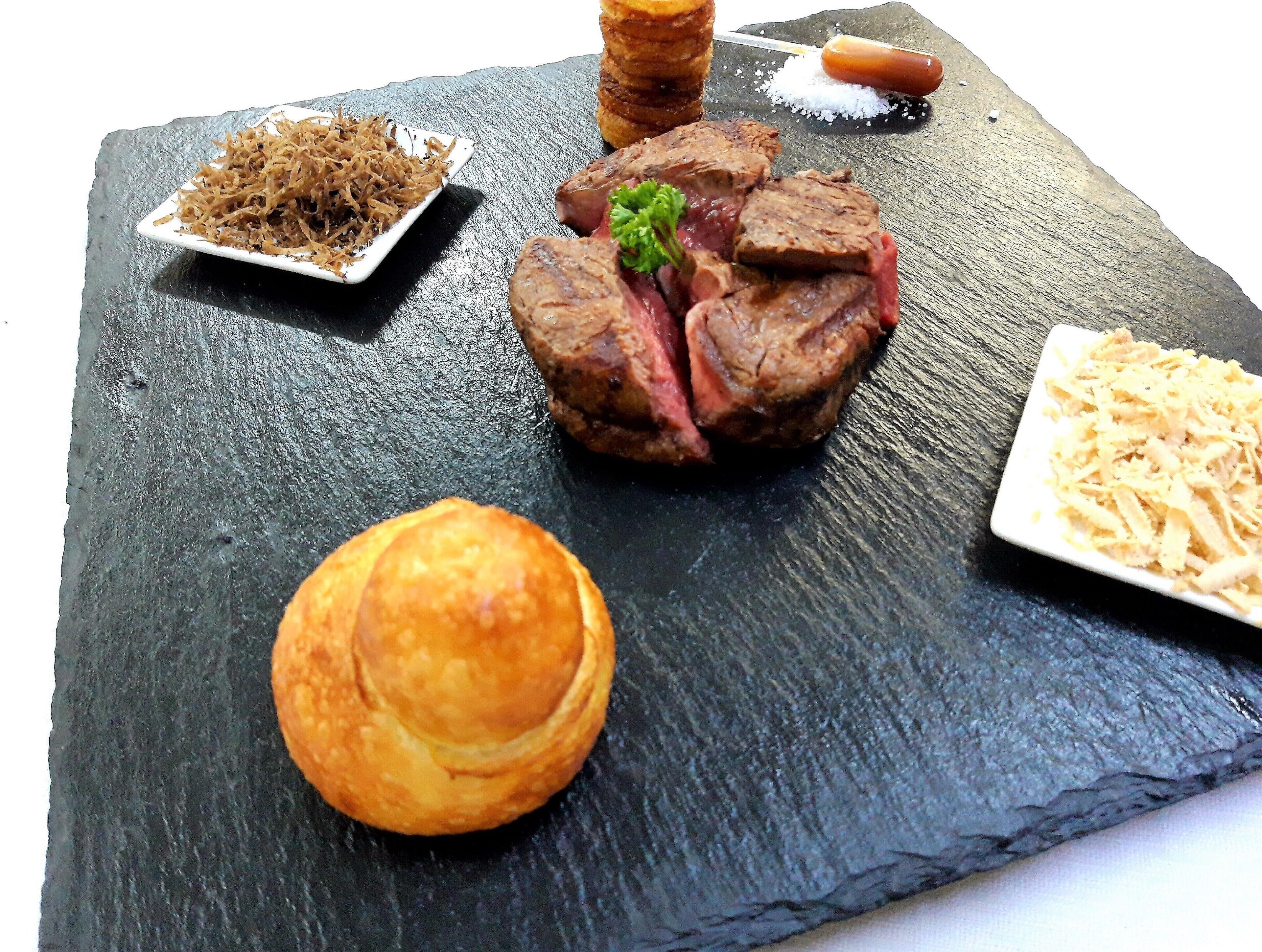 Eine schwarze Schiefer Servierplatte mit einem Steak, einer kleinen Schüssel geriebenem Käse und einem runden Gebäckstück.