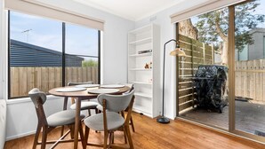 Un coin repas avec une table ronde et des chaises, une étagère blanche et une vue sur le jardin à travers une porte-fenêtre coulissante.