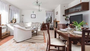 Een woonkamer met een bank, salontafel, eettafel gedekt voor vier personen en een aan de muur bevestigde televisie.