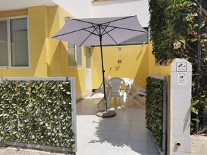 Ein gelbes Gebäude mit einer weißen Tür, einem Überwachungsgehäuse und einer Terrassenfläche mit Tisch und Stühlen unter einem großen Sonnenschirm.