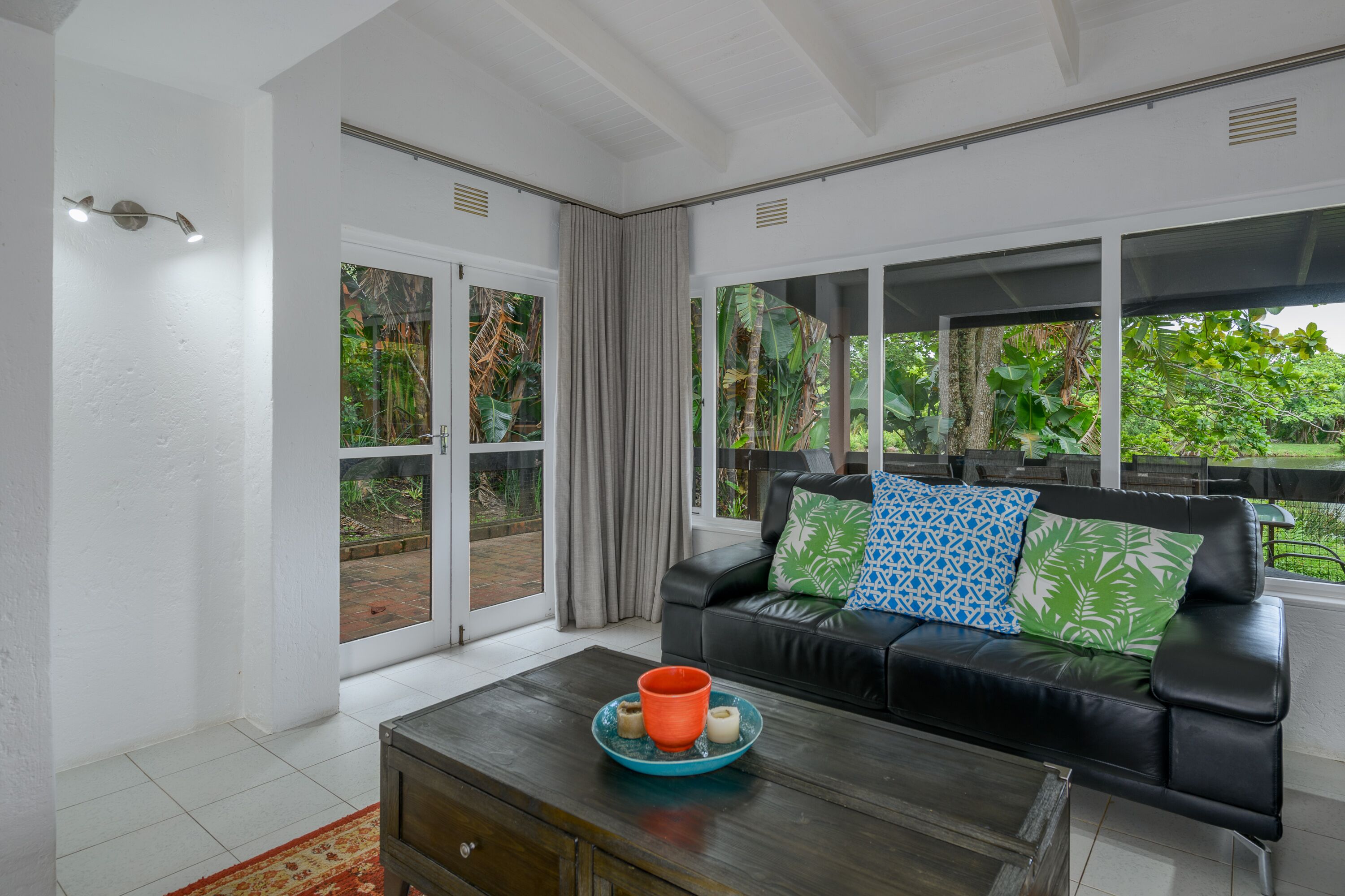 A modern living room with a black leather sofa, a coffee table with a blue tray, and a view of greenery through large windows.