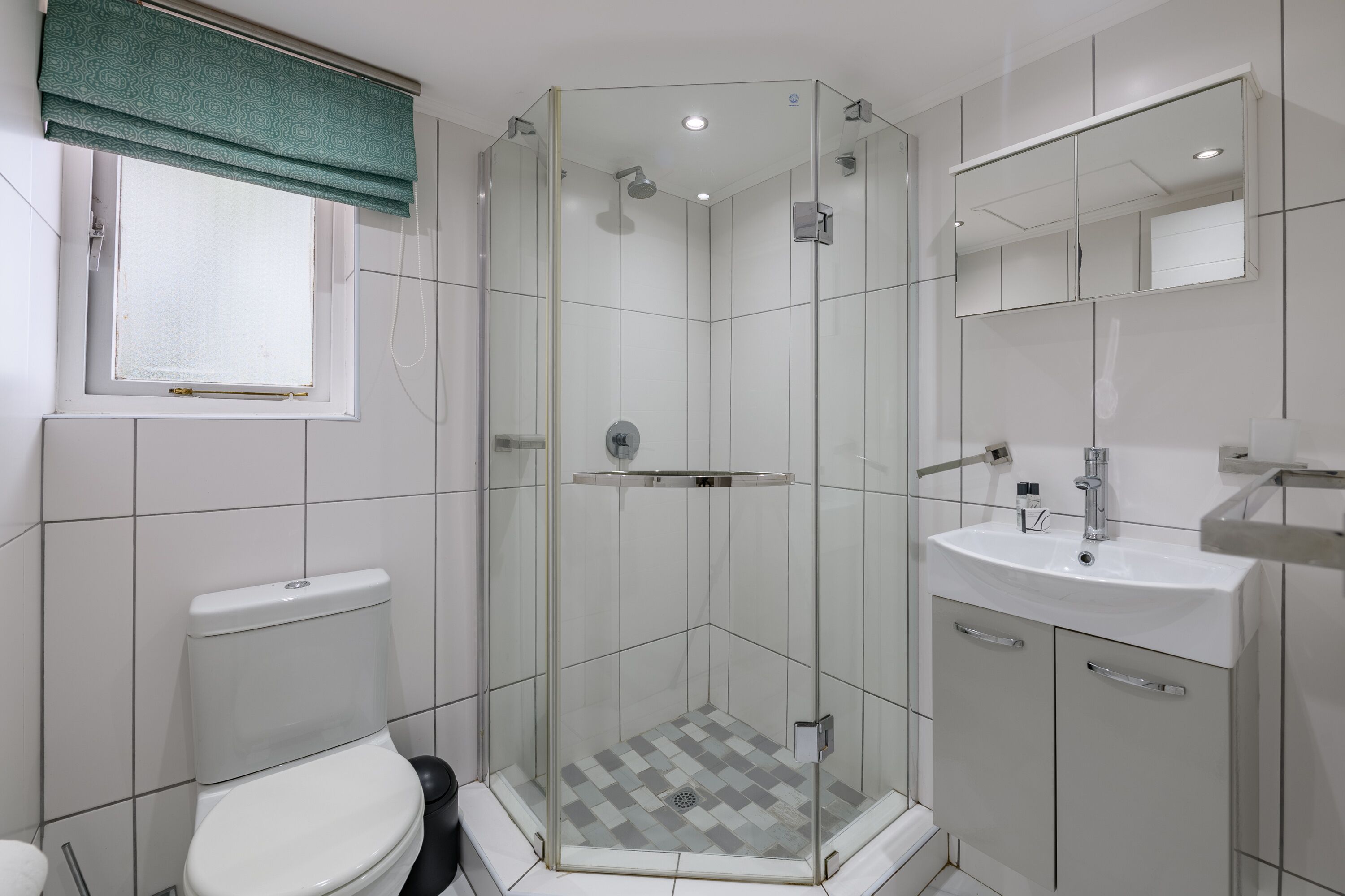 A modern bathroom with a glass-enclosed shower, a toilet, a sink with a mirror, and a window with a green curtain.