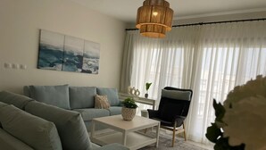 A living room with a sofa, a coffee table, a chair, a hanging light fixture, and a window with sheer curtains. - Marassi Marina 2 bedroom pool view 403 (El Alamein)